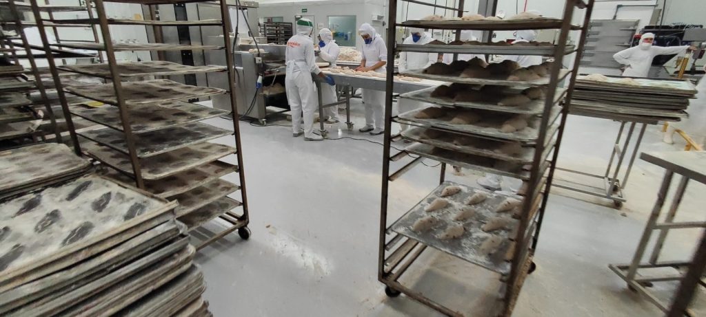 Linha de produção de panificação com piso epóxi cinza, funcionários manipulando massas e carrinhos com assadeiras.