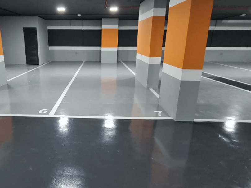 Estacionamento interno com piso epóxi cinza e áreas demarcadas em branco, colunas com faixas decorativas laranja e cinza.