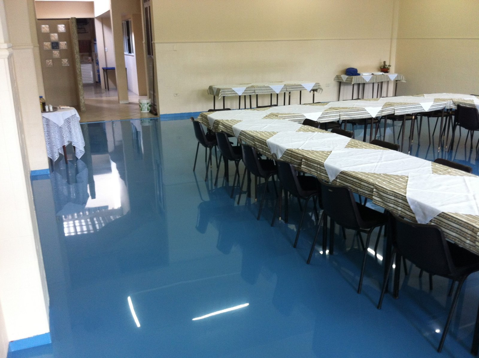 Salão com piso epóxi azul brilhante refletindo a luz, mesas dispostas para refeição ou evento.