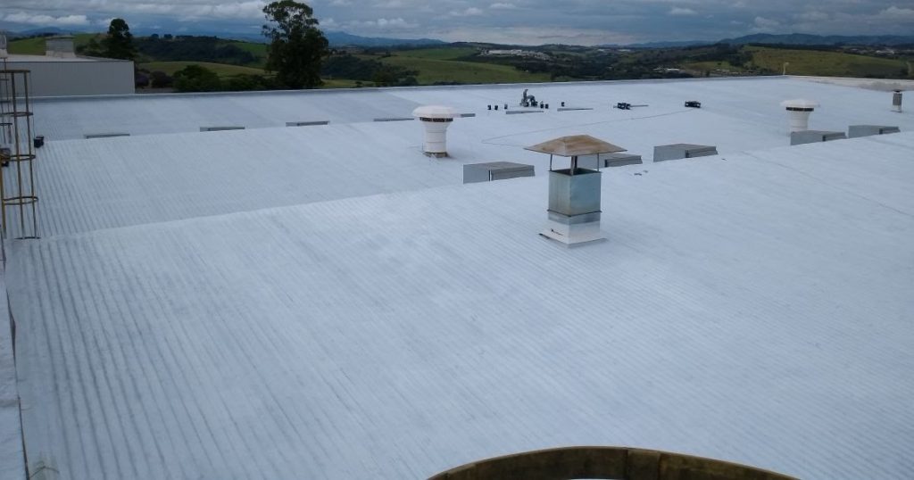 Cobertura industrial ampla com aplicação de impermeabilização branca e vista de paisagem ao fundo.