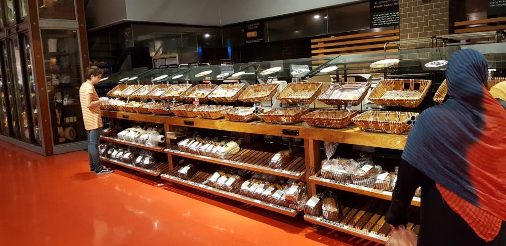 Setor de padaria de supermercado com piso epóxi vermelho, prateleiras de madeira e cestos de pães vazios.